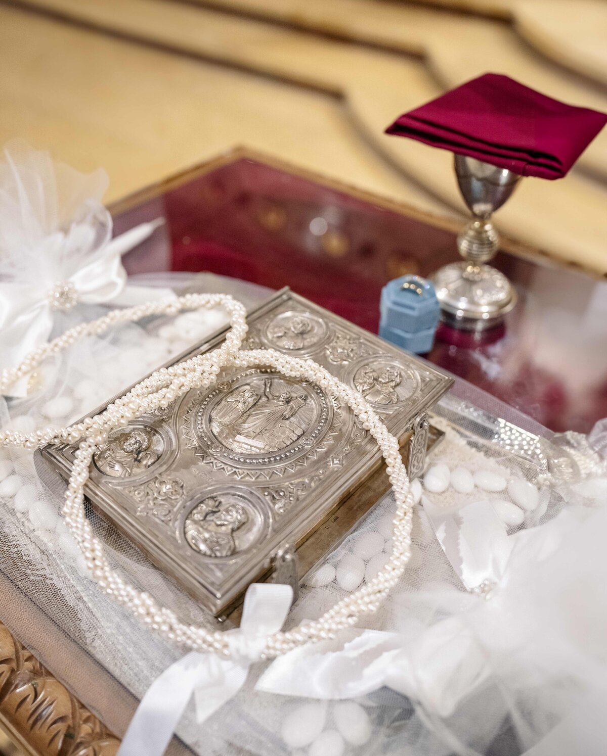 A decorative box with religious engravings is placed on a glass table. A string of pearls is draped over it. Nearby are a silver chalice with a folded red cloth and a blue candle holder, all arranged on a white lace cloth.