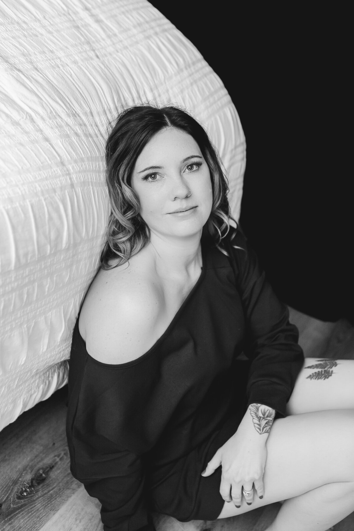 A woman sitting on the floor next to the bed, wearing a navy blue top, her tattoo visible, exuding a sense of quiet reflection.