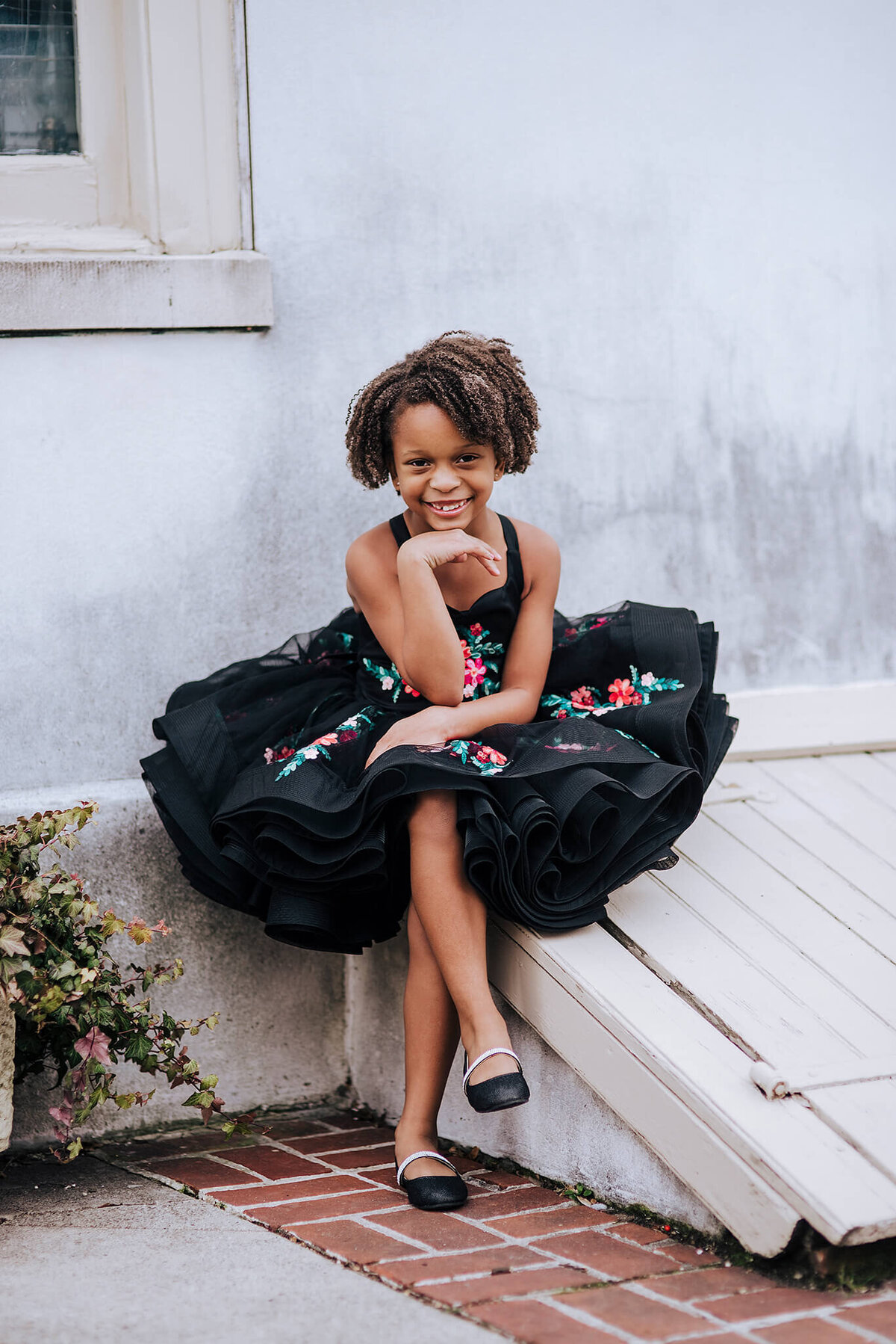 African American girl in a black floral dress with black shoes smiling with chin on her hand sitting on a city street in Fells Point Baltimore Maryland