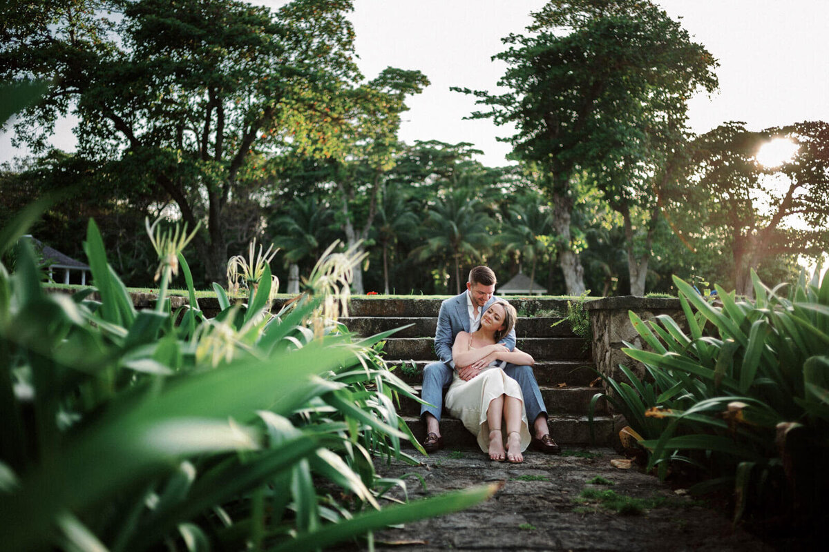 The engaged couple is sitting on a stone staircase in the middle of lush greens in Round Hill Hotel and Villas, Jamaica.