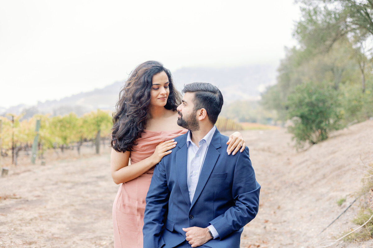 man in a blue suit sits and looks over his shoulder at his fiance who is in a coral pink dress holding his shoulders for their engagement pictures by bay area photographers