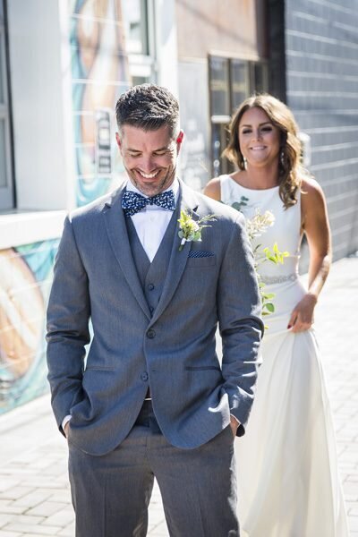A groom smiles down at the ground as his bride approaches from behind for their first look.