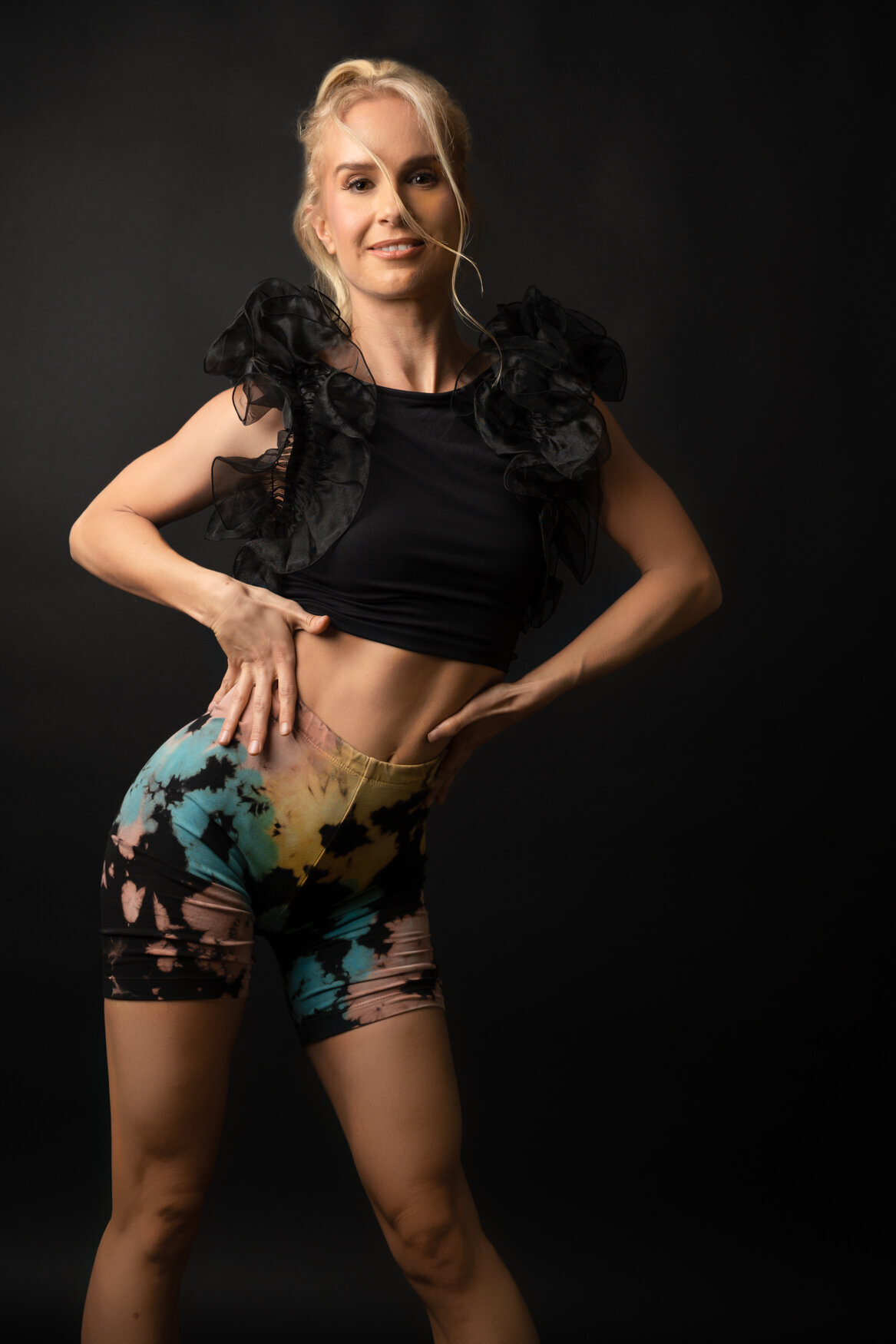 Stylish portrait of a woman against a sleek black background. She is wearing a chic black top with ruffled detailing and vibrant tie-dye shorts, striking a confident pose that exudes elegance and creativity