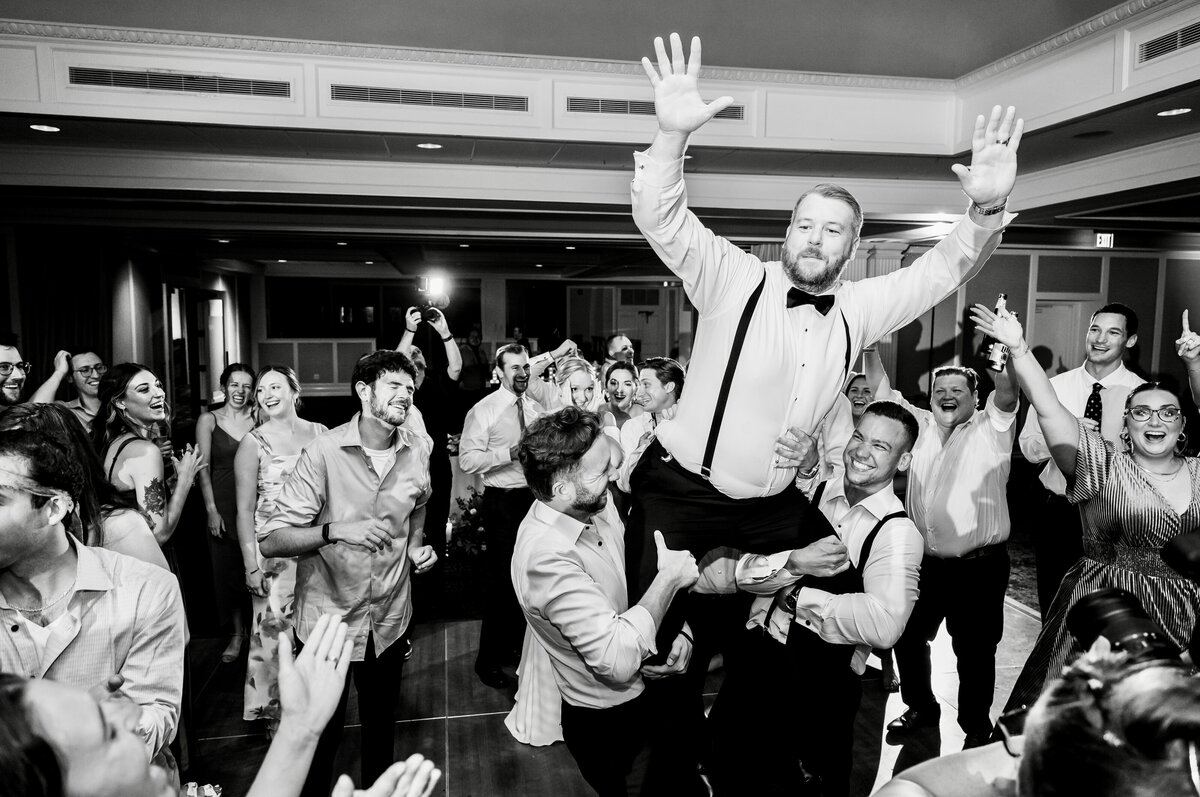 Black and white photo of a man in a bow tie being lifted in the air by others at a lively party, surrounded by smiling and cheering people.