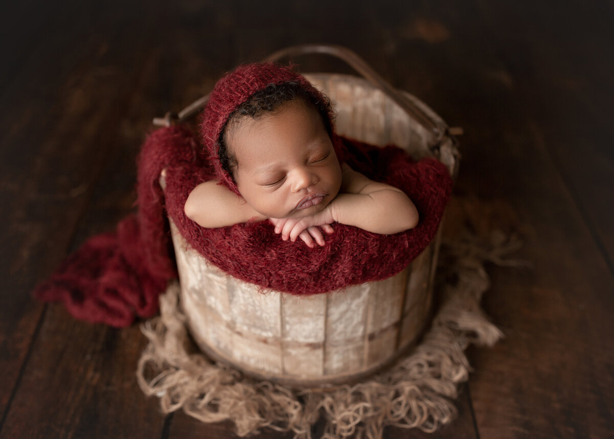 Baby sleeping in a bucket for newborn photoshoot. Baby's hands are folded under his chin and his cheek is resting on his left arm. Baby is sleeping.