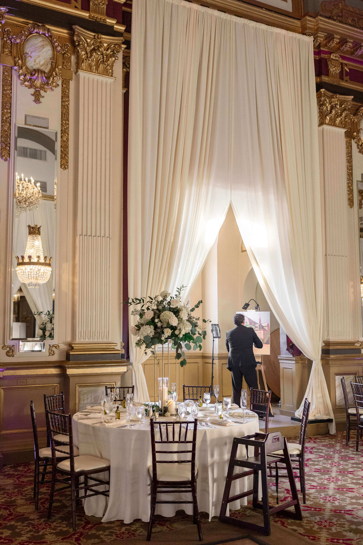 An elegant ballroom with tall white drapes and ornate gold details. A formally dressed man stands in front of a curtain-covered stage, while a round table with a white cloth and floral centerpiece is set with chairs on a patterned carpet.