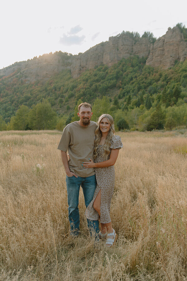 A husband and wife smile for a couples photo portrait. They're outdoors with the eastern Idaho landscape behind them.