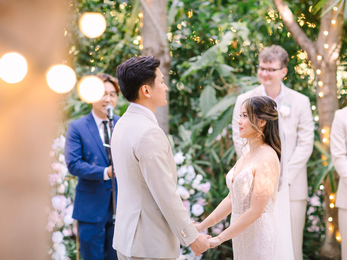 A couple in elegant attire holds hands, exchanging vows outdoors under soft light bulbs and lush greenery.