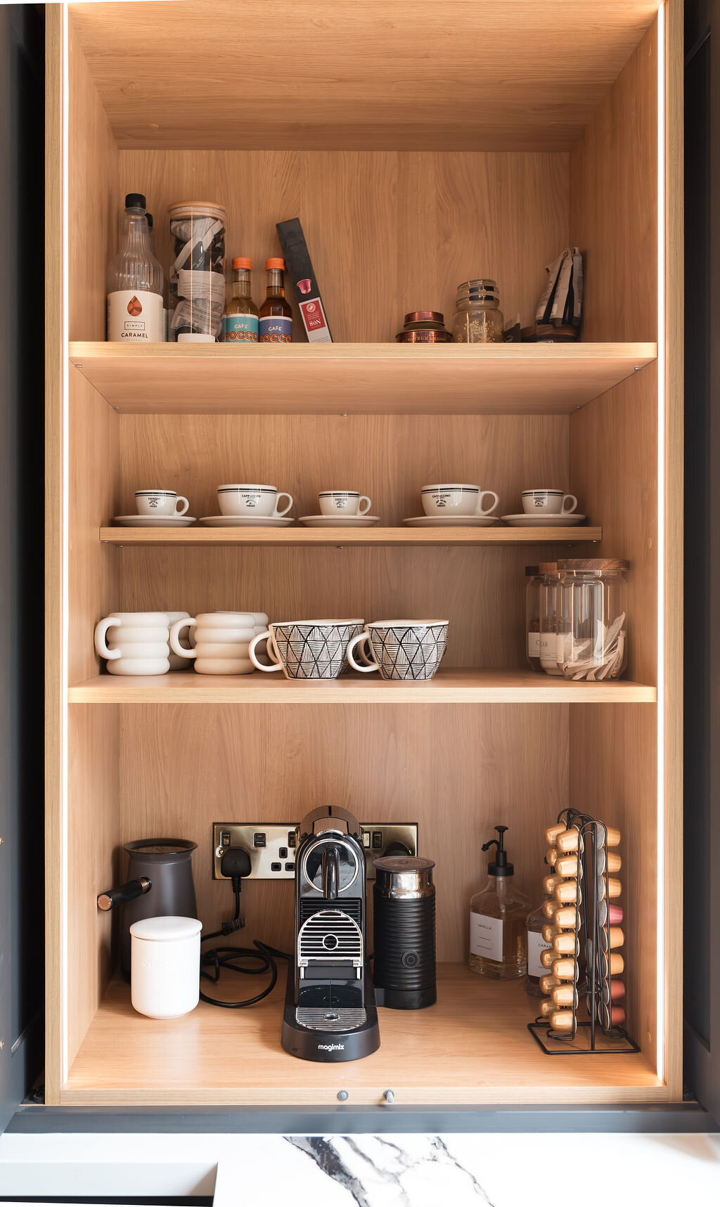 A built-in kitchen cabinet with black exterior and wood interior, illuminated by built-in lighting, displays shelves with mugs, glasses, spices, and coffee supplies. The counter features a coffee machine, capsule holder, and various bottles.