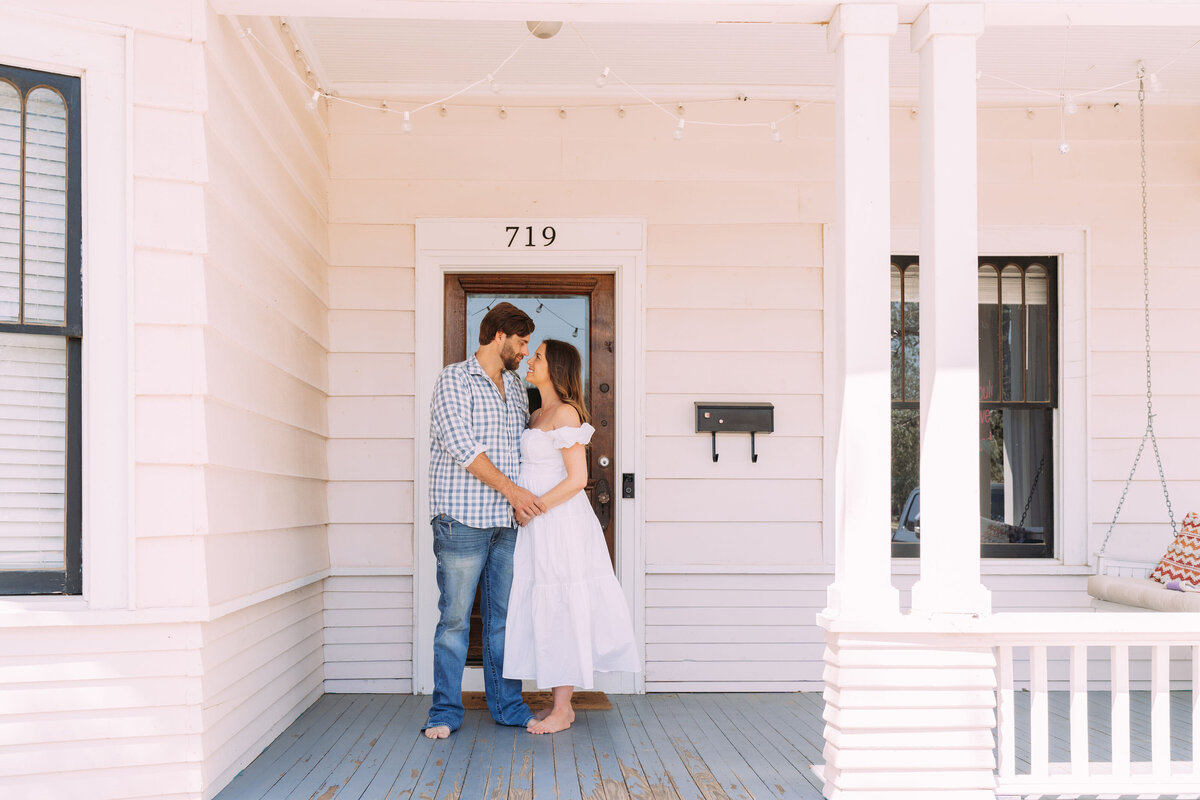 expectant parents stand on front porch with mother in white dress
