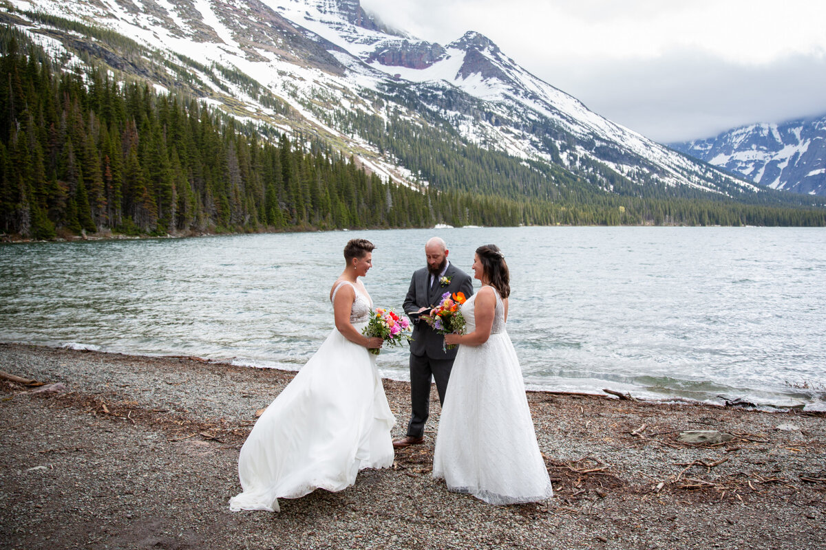 Two brides stand facing each other, holding bouquets and listening as their officiant reads the ceremony script. There are snow capped mountains and a lake behind them.