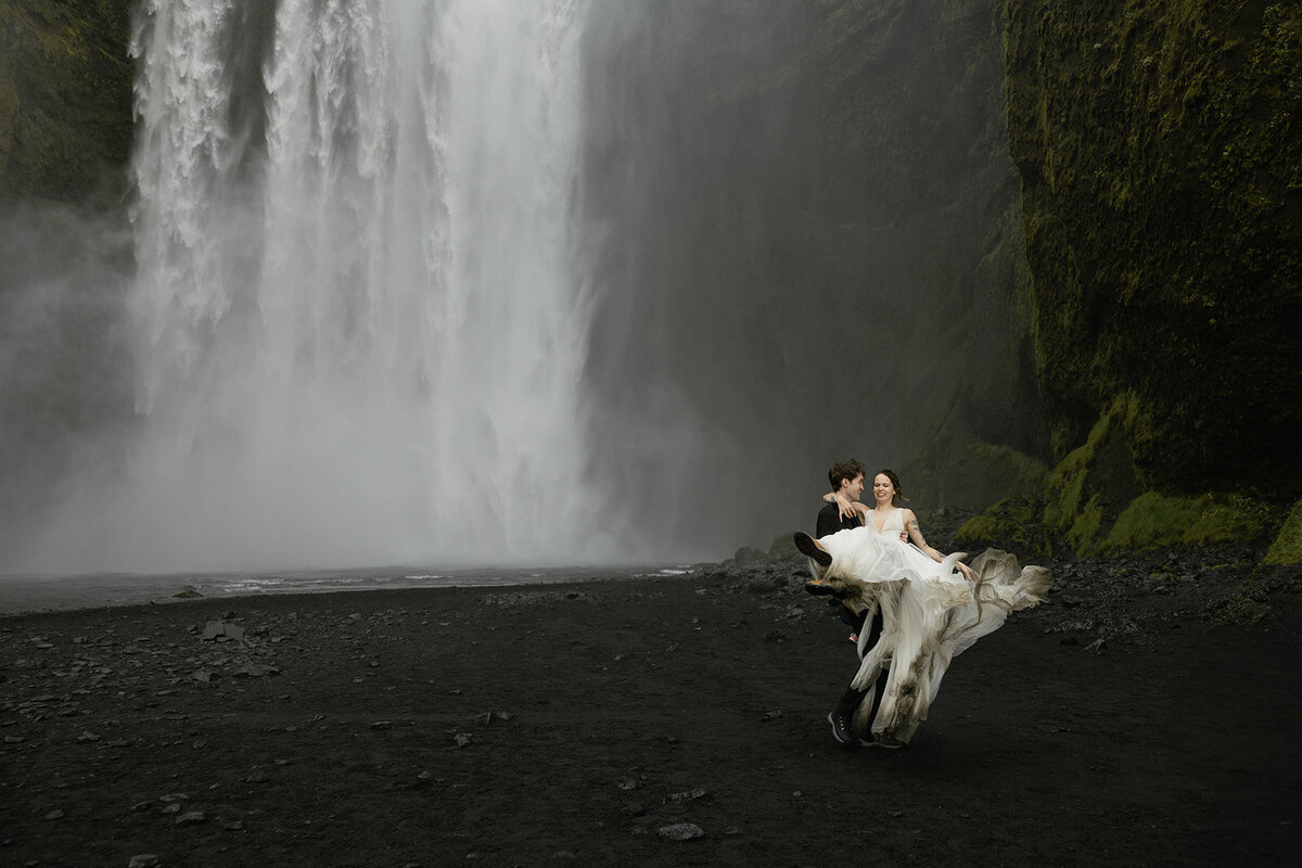 Groom lifting bride in front of the powerful Skógafoss waterfall in Iceland during their elopement, captured by a destination elopement photographer.