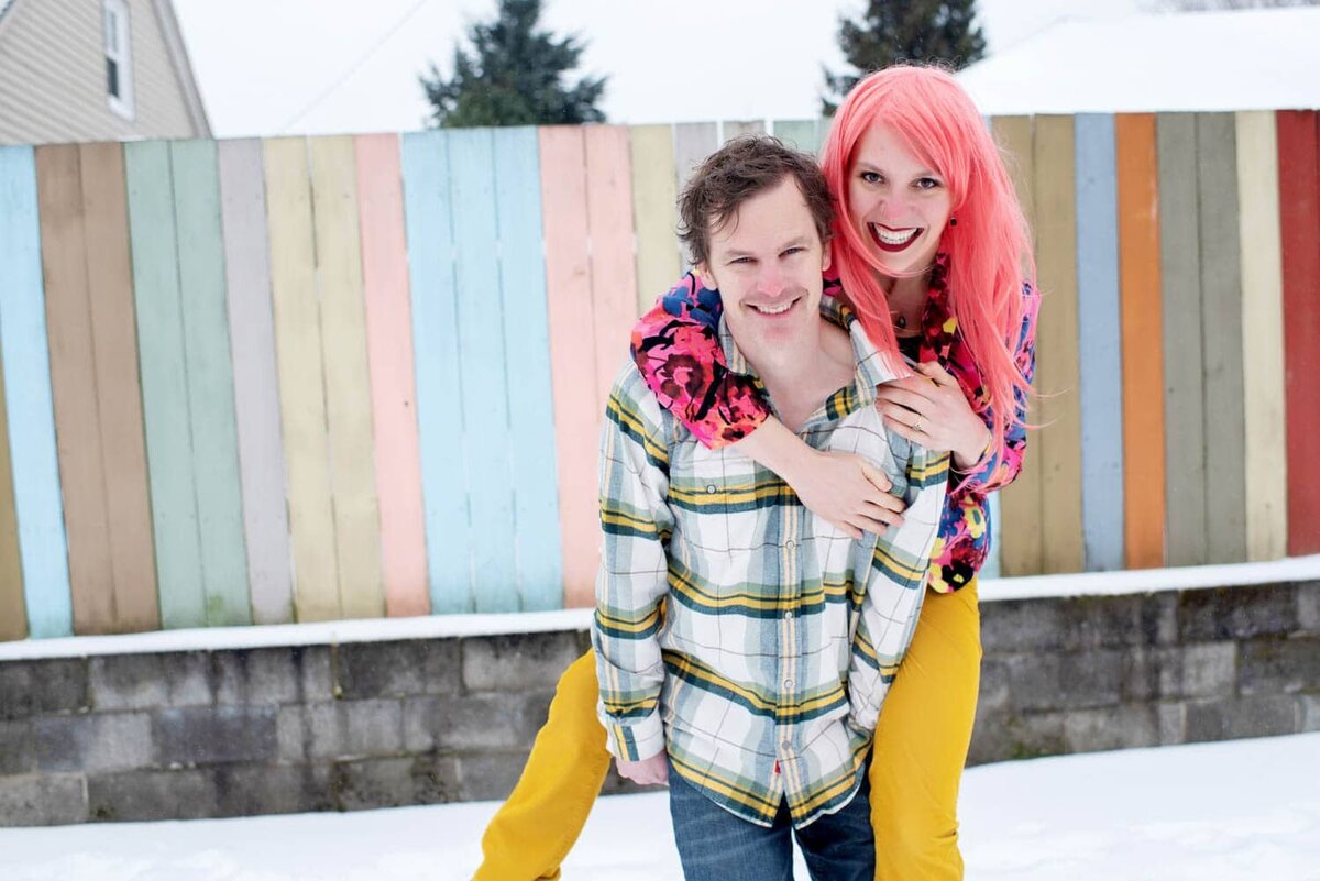 a woman with bright pink hair jumps on her fiance's back in front of a colorful fence during engagement photos in portland