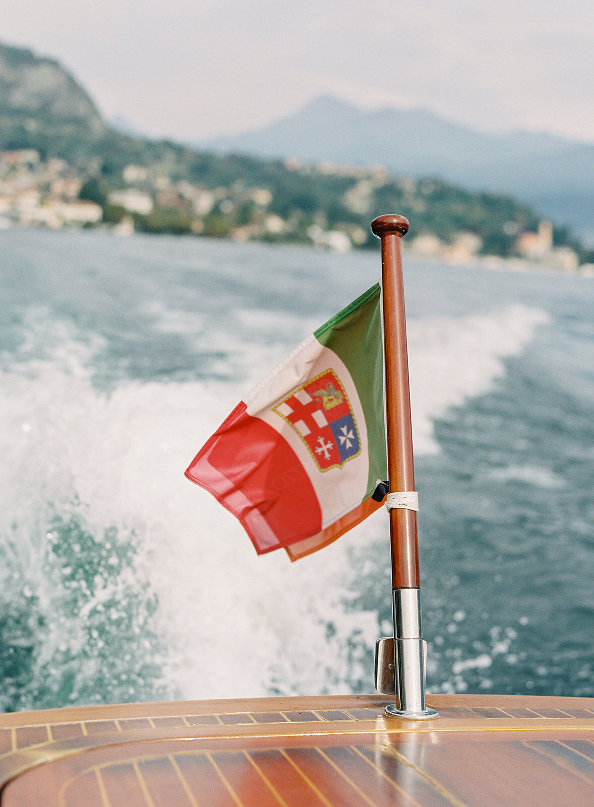 Film photograph of bride and groom on a wooden boat on Lake Como in Italy she is wearing a white halter jumpsuit with her hair down in loose waves and he is wearing grey and navy cashmere sweater and coat photographed by Italy wedding photographer