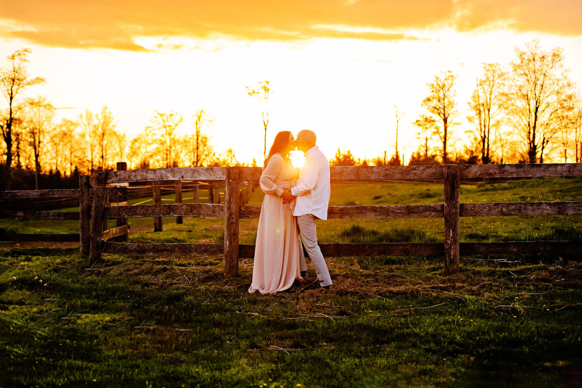 husband and wife standing on a farm with a wood fence and the sun setting behind for their outdoor summer maternity photography session in Burlington, On