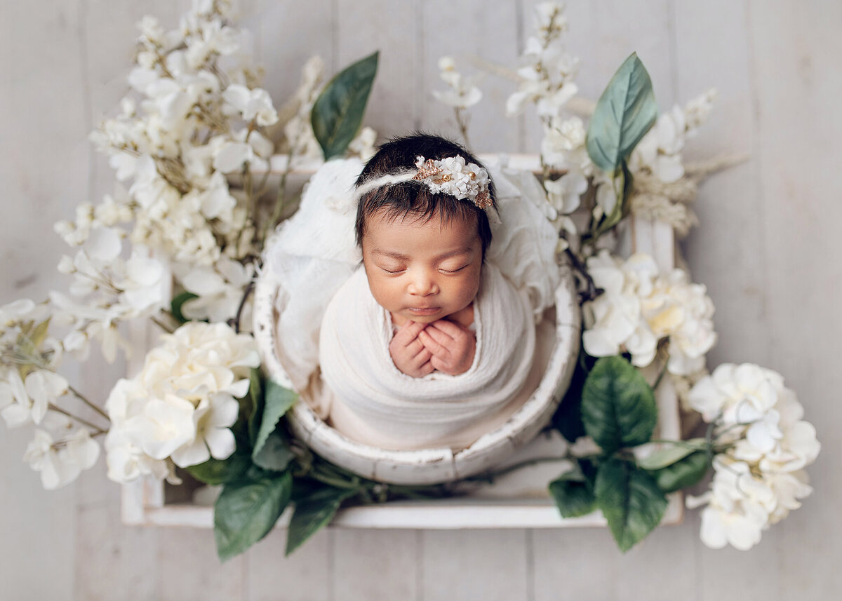 Little baby girl with lots of dark hair snuggled in a wrap and a bucket surrounded by large white flowers