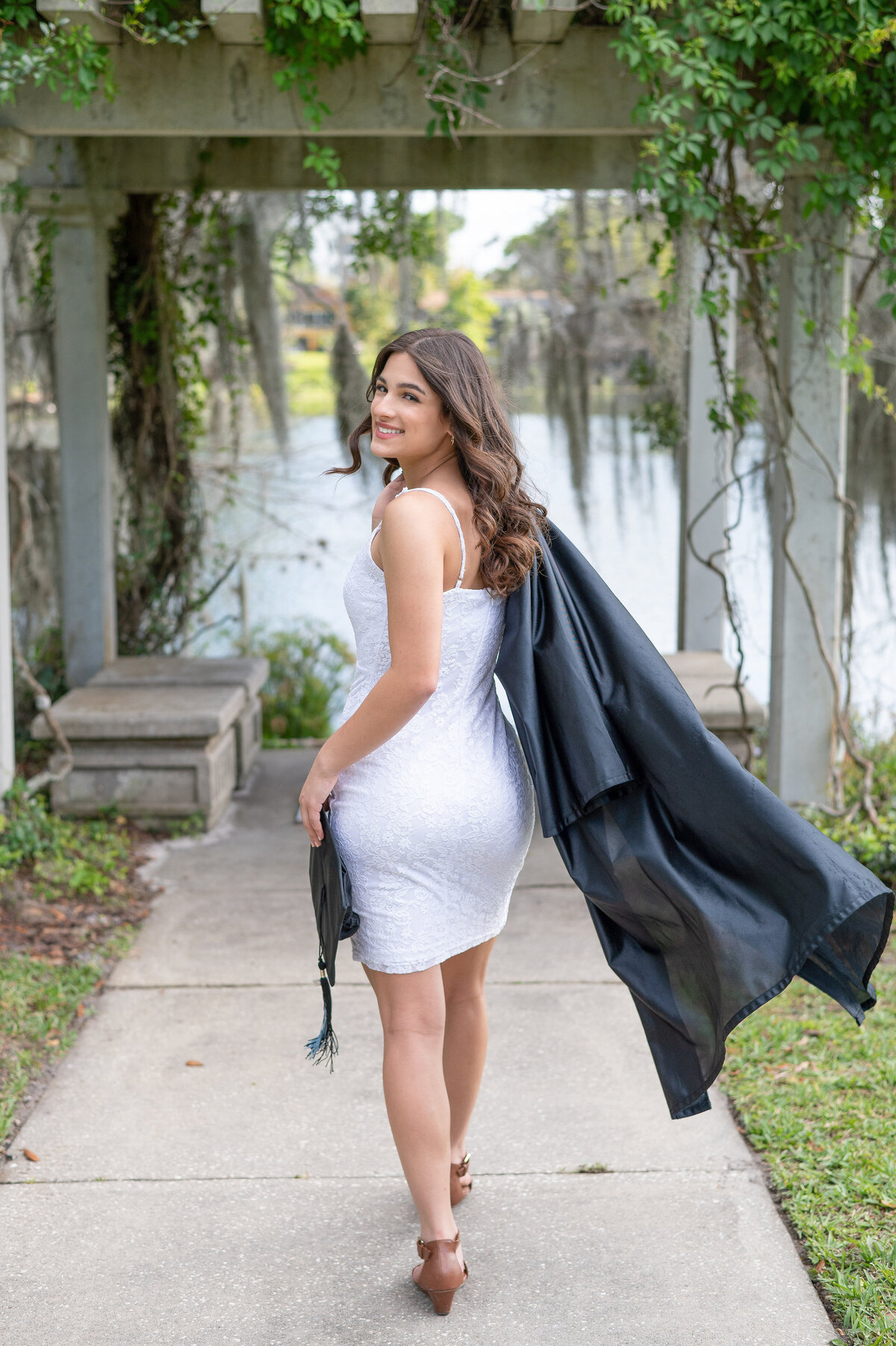 High school senior girl holding cap and gown looks back at camera smiling.