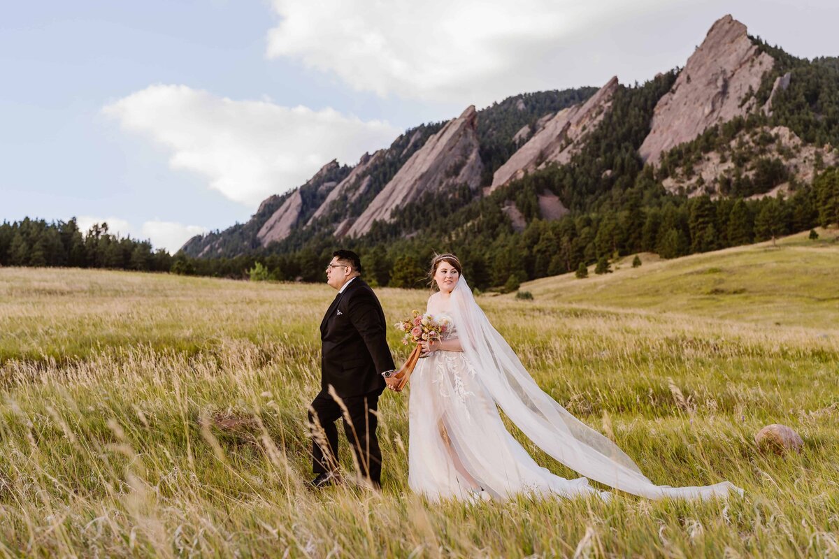 Bride and groom walk hand in hand through a golden field with the Flatirons in the background during their Boulder, Colorado elopement.