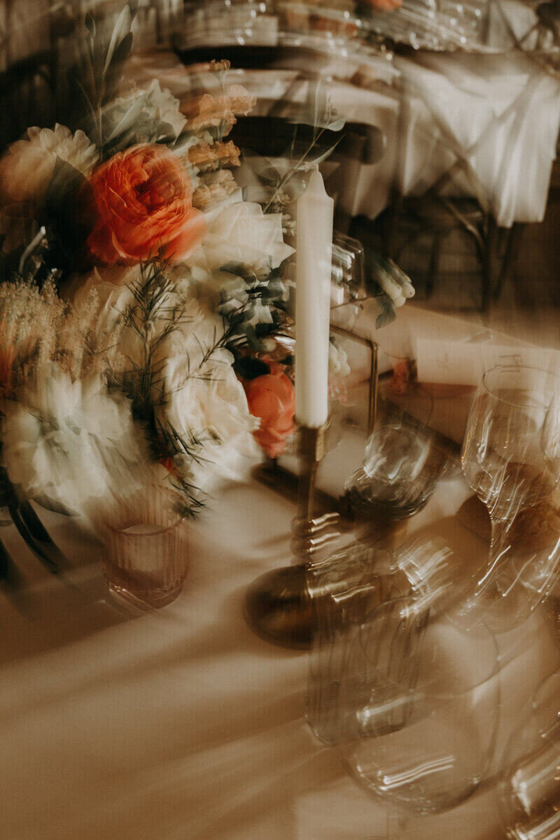 Bougeoir doré surmonté d'une bougie blanche, verres et bouquet de fleur formant un tourbillon flou. Scène capturée par Laura, photographe de mariage en vendée.