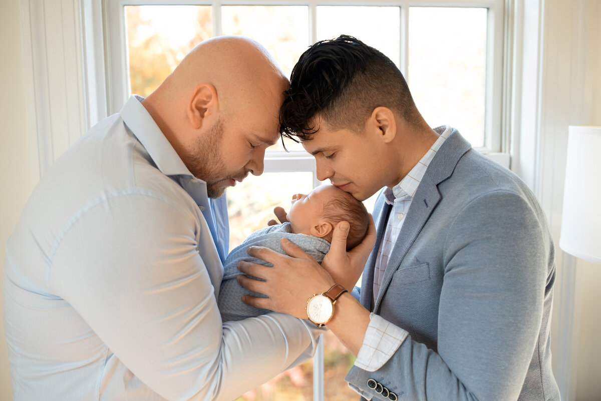 two dads and their newborn baby boy in home session dads wearing suits in front of window touching foreheads and looking at baby with soft blues and creams