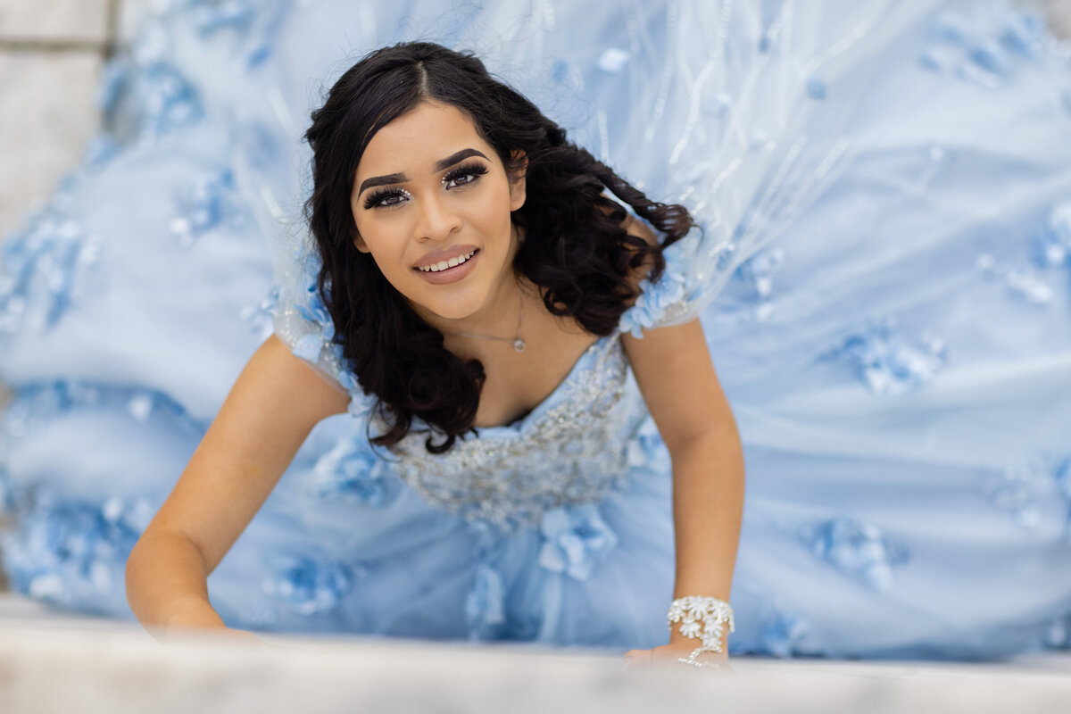 A joyful close-up of Victoria lying gracefully in her stunning blue quinceañera gown, captured during her photoshoot at University Circle in Cleveland, Ohio. TheWiz Photography highlights her beauty and happiness on this special day.