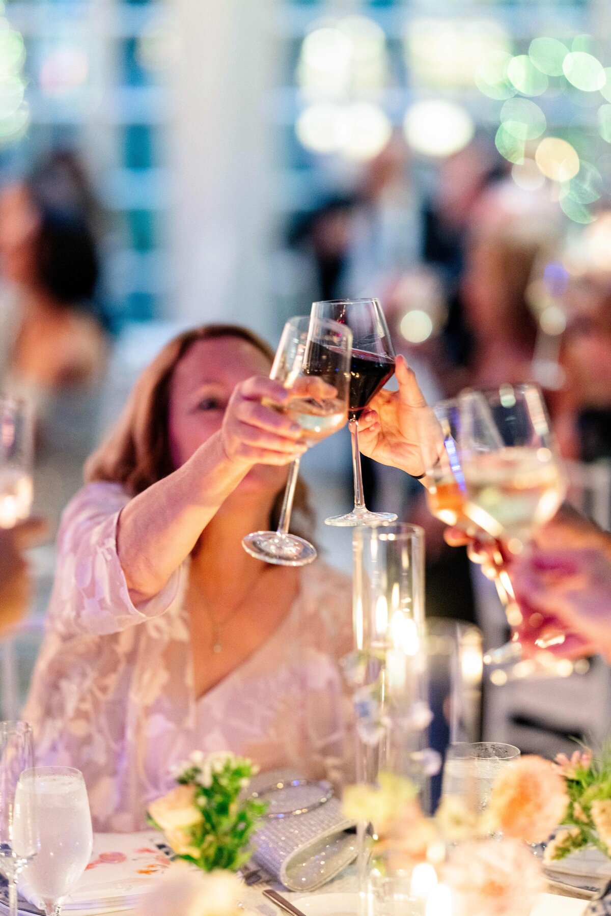 People clink wine glasses in a celebratory toast at a dining table adorned with lit candles and floral arrangements. The setting is warm and festive, with blurred lights in the background creating a joyful ambiance.