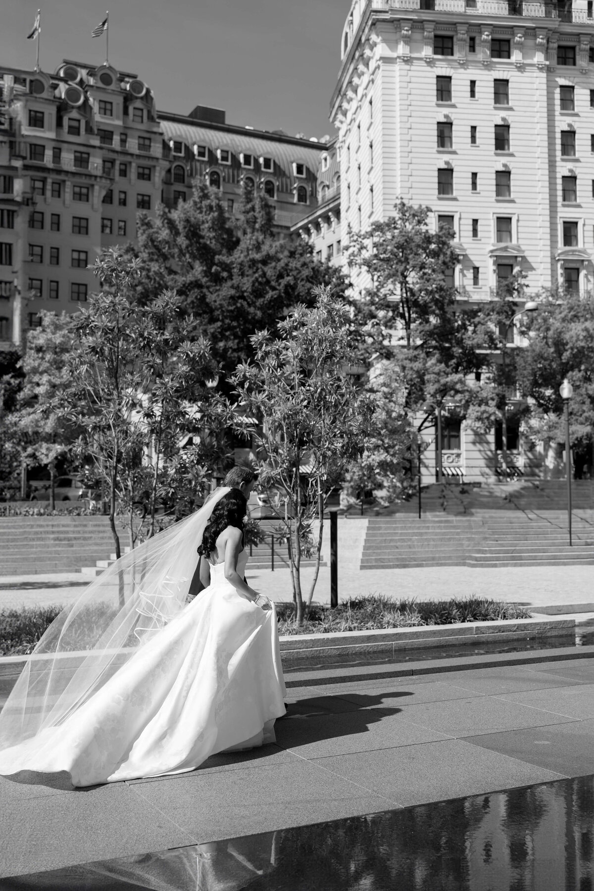 A bride in a long white gown and veil walks outside in an urban setting, surrounded by trees and large buildings, on a sunny day. The scene is in black and white.