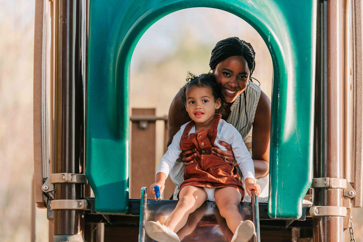 Little girl sitting on a slide with her mom behind her. 