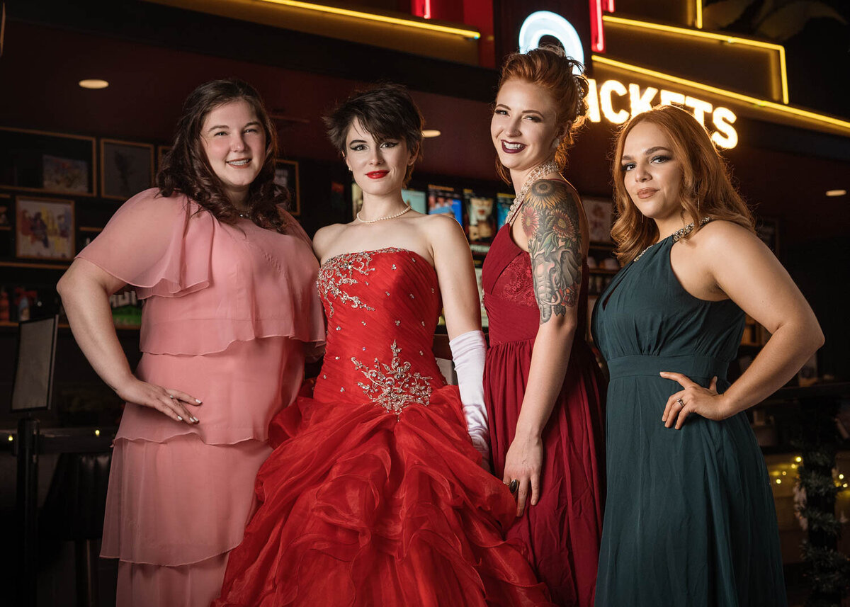 Group portrait of four women dressed in elegant gowns, smiling and posing in front of a movie concession stand backdrop.