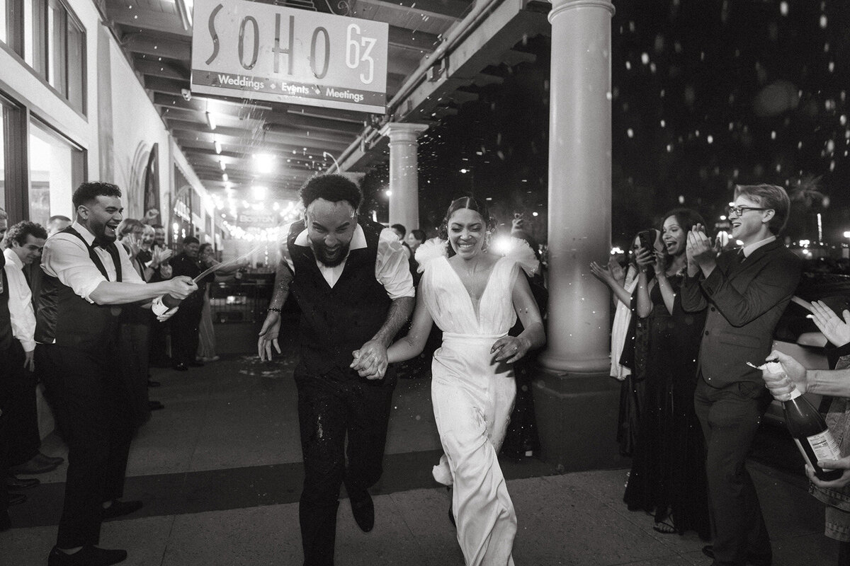 A joyful bride and groom run hand-in-hand through a tunnel of cheering guests outside SoHo63, a popular Chandler wedding and event venue. The black-and-white photo captures the couple smiling as friends and family celebrate around them under a covered outdoor walkway.