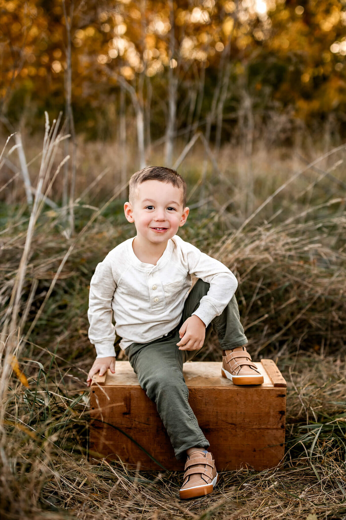 A young boy with short hair is perched on a wooden crate in a grassy field, perfect for outdoor portraits. He wears a white henley shirt, green pants, and brown shoes. Behind him, the tall grass and trees are bathed in a warm, golden hue that any Bloomington family photographer would cherish capturing.
