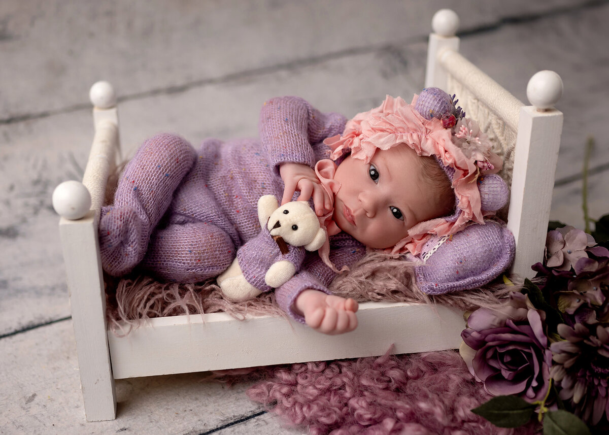 A newborn in a purple onesie cuddles a tiny bear in a small wooden bed