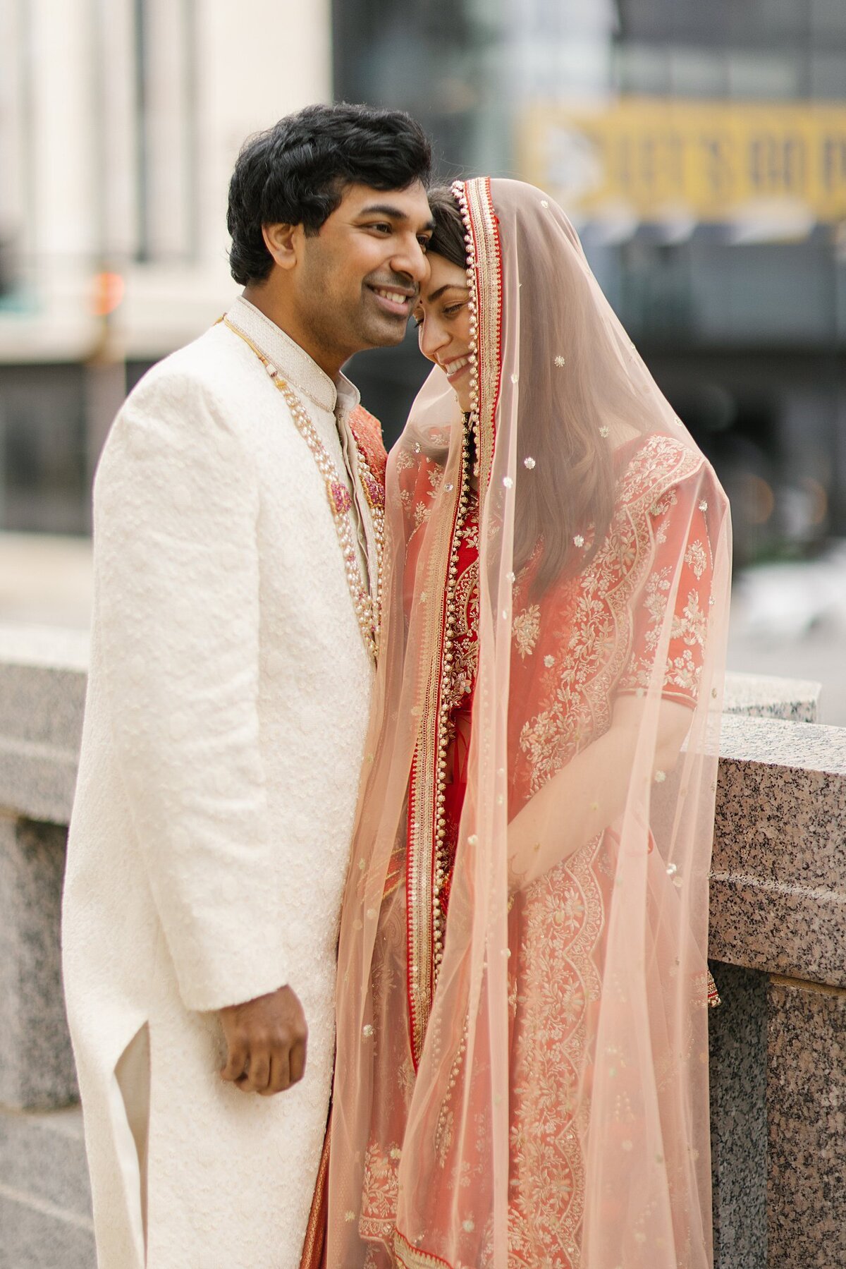 Indian bride in a red and gold wedding saree embraces the Hindu groom wearing a white sharwani with a gold wedding necklace outside the Frist Museum of Art in Nashville, TN