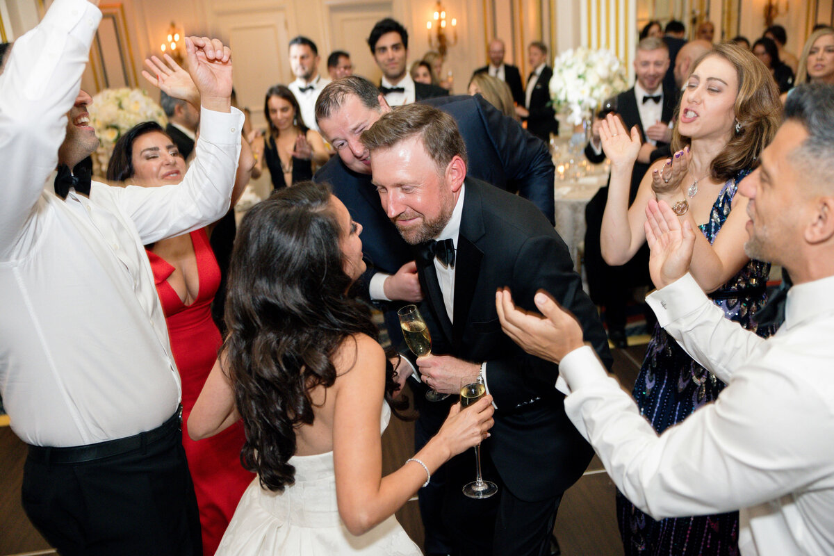 A joyful wedding celebration with a bride and groom dancing among guests. People are clapping and cheering, creating a lively atmosphere. The guests are dressed in formal attire, surrounded by elegant decor.