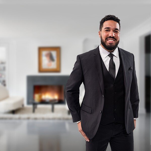 A happy man dressed in a black wedding suit poses with a fireplace in the background