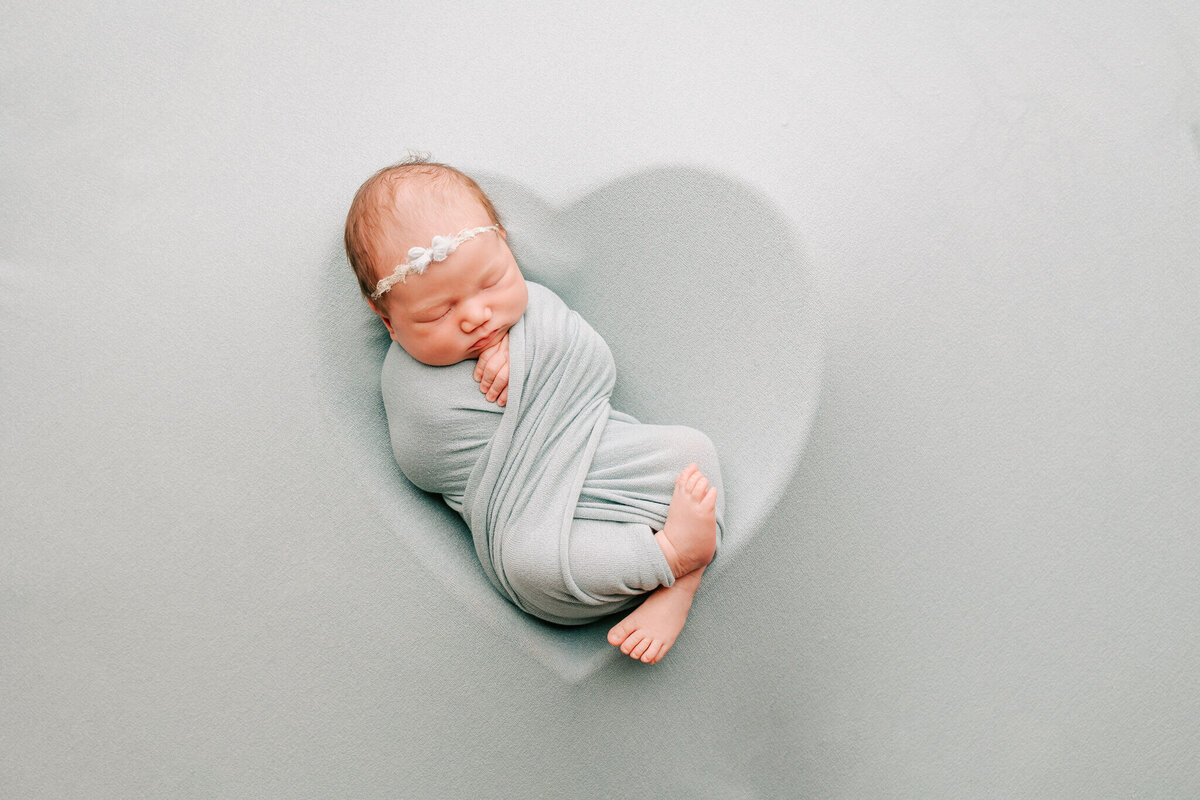sweet portrait of beautiful newborn baby girl who is wrapped in teal wrap laying on teal heart outline.