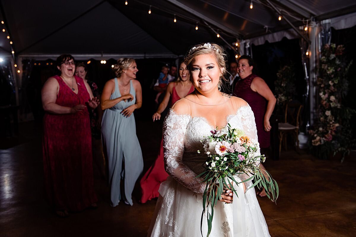 Bridesmaids and wedding guests dressed in burgundy and light blue line up behind the bride wearing a long sleeved lace wedding dress with an off the shoulder plunging neckline as she throws her bridal bouquet of  burgundy and blush pink flowers and greenery at Arrington Vineyards.