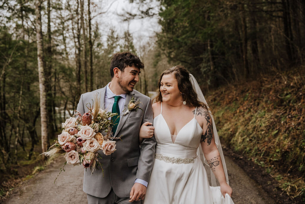 Newlywed couple walking arm in arm down a forest road near Greenbrier, holding a bouquet after their Gatlinburg elopement.
