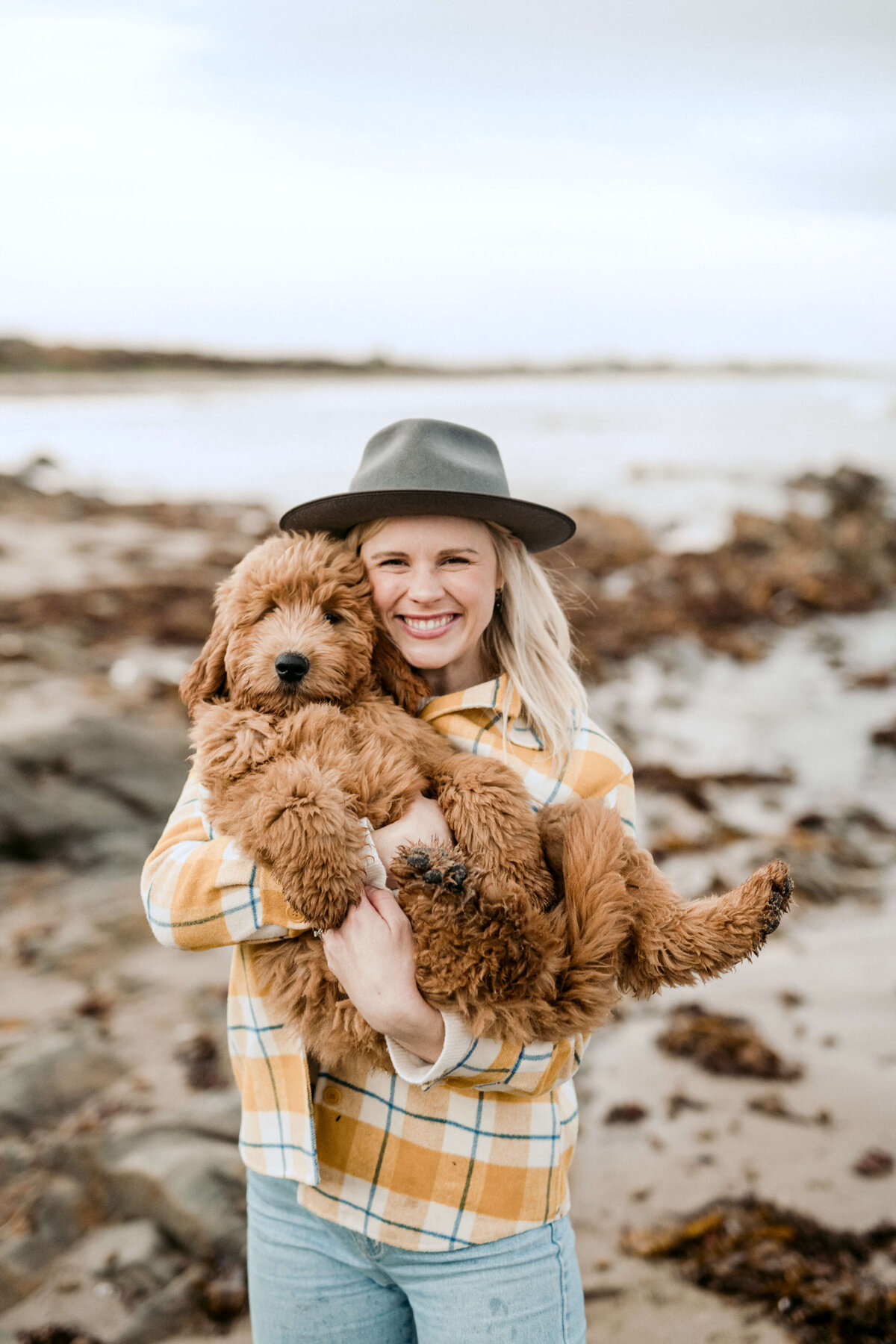 Jessie and her dog Eddie on the beach at sunset, both looking at the camera and smiling. Jessie wears a hat and winter clothing.