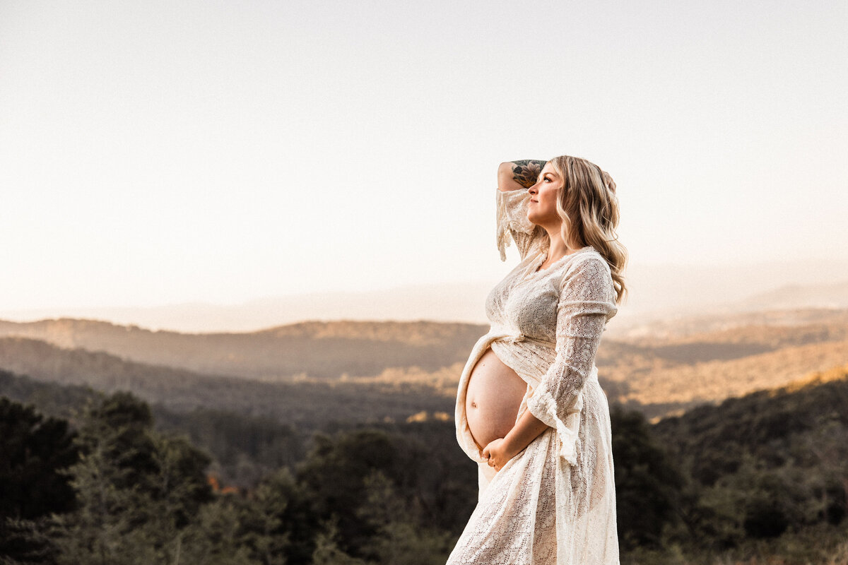 Pregnant woman stands against hills backdrop while leaning back and holding belly