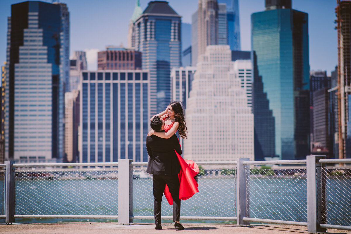 groom picking up the bride in red formal outfit during their engagement shoot at the brooklyn bridge park in dumbo nyc