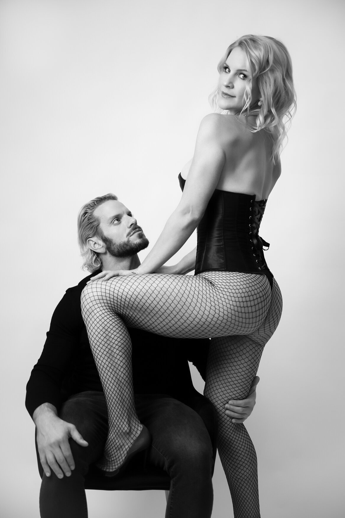 Black and white portrait of a couple in an intimate pose. The woman, dressed in a corset and fishnet stockings, sits confidently on the man's lap, looking toward the camera. The man gazes up at her with admiration, one hand resting gently on her leg.
