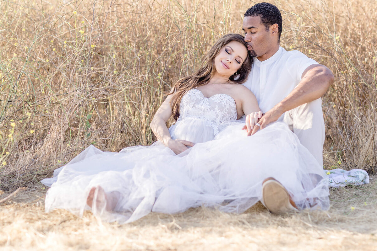 Husband and wife dressed in all white sitting in a field of tall brown grass. Husband kissing wife's head while she leans on his shoulder, cradling her baby bump with her eyes closed.