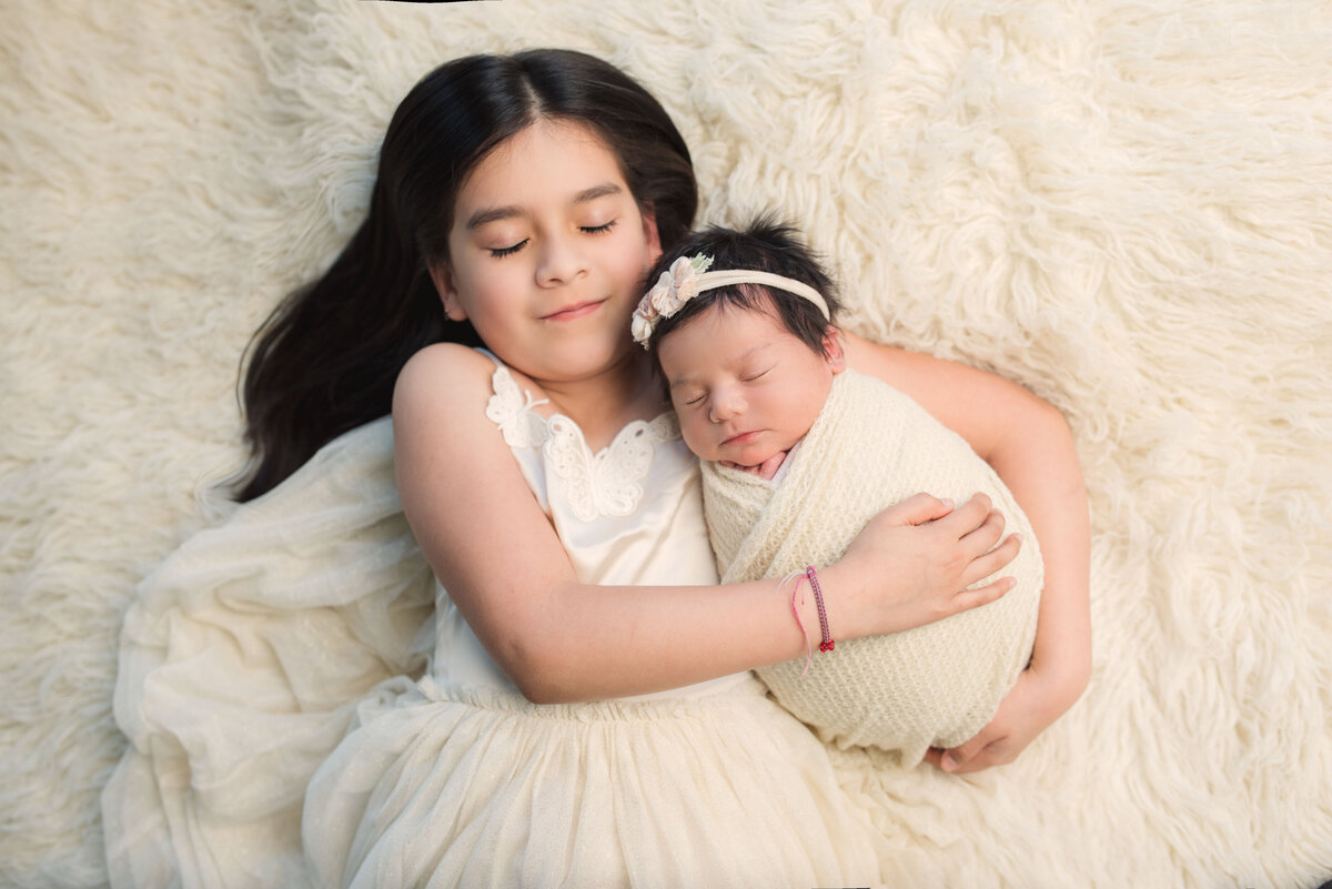 Big sister holding newborn sister while lying down on the white fur rug, both are wearing white and their eyes are closed
