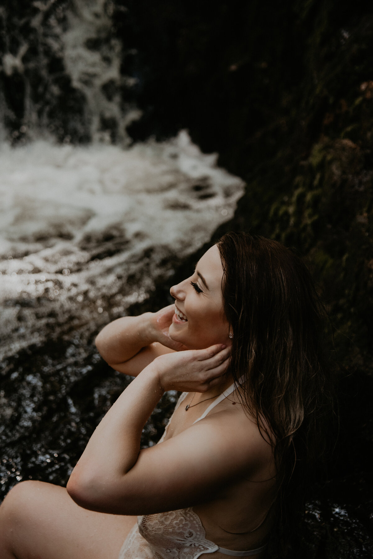 side view of a girl sitting on a boulder beside a waterfall. She has her hands up around her jawline and she is laughing. She is wearing a white body suit and has wet hair from being in the small waterfall catch.