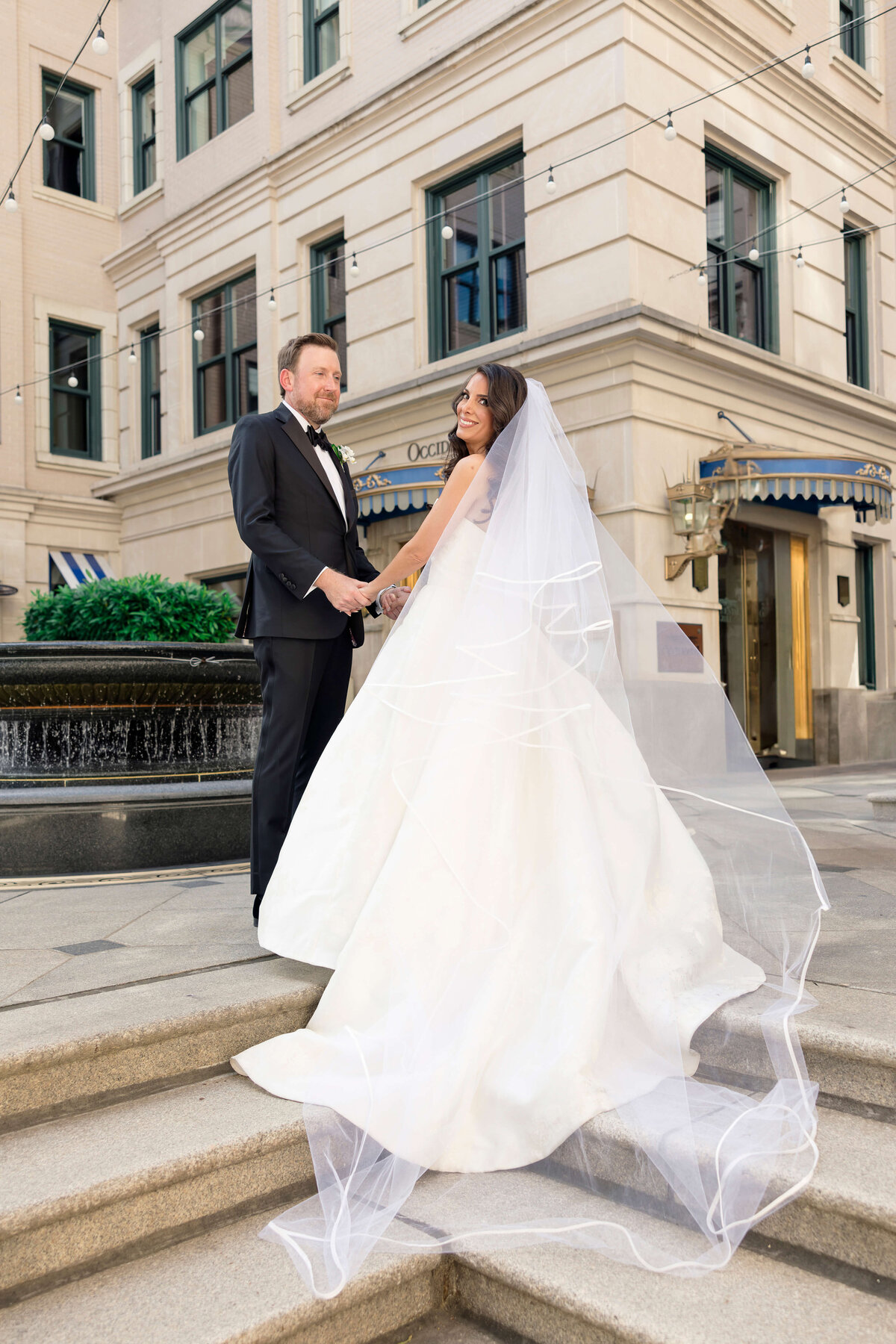 A bride in a white gown with a long veil and a groom in a black suit stand holding hands on stone steps in front of a building. They are smiling at each other, with string lights overhead and a fountain nearby.