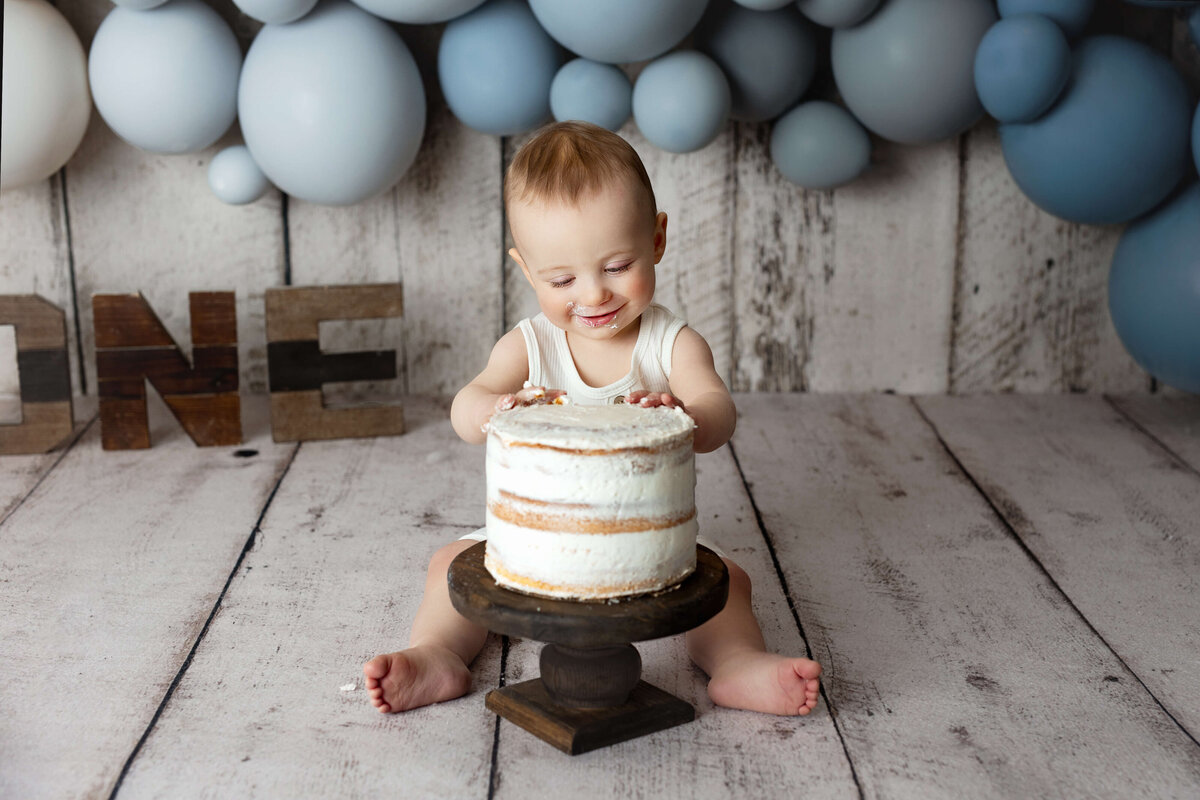 1 year old boy eating cake on a white wood background with blue balloons and ones letters during a cake smash photography session