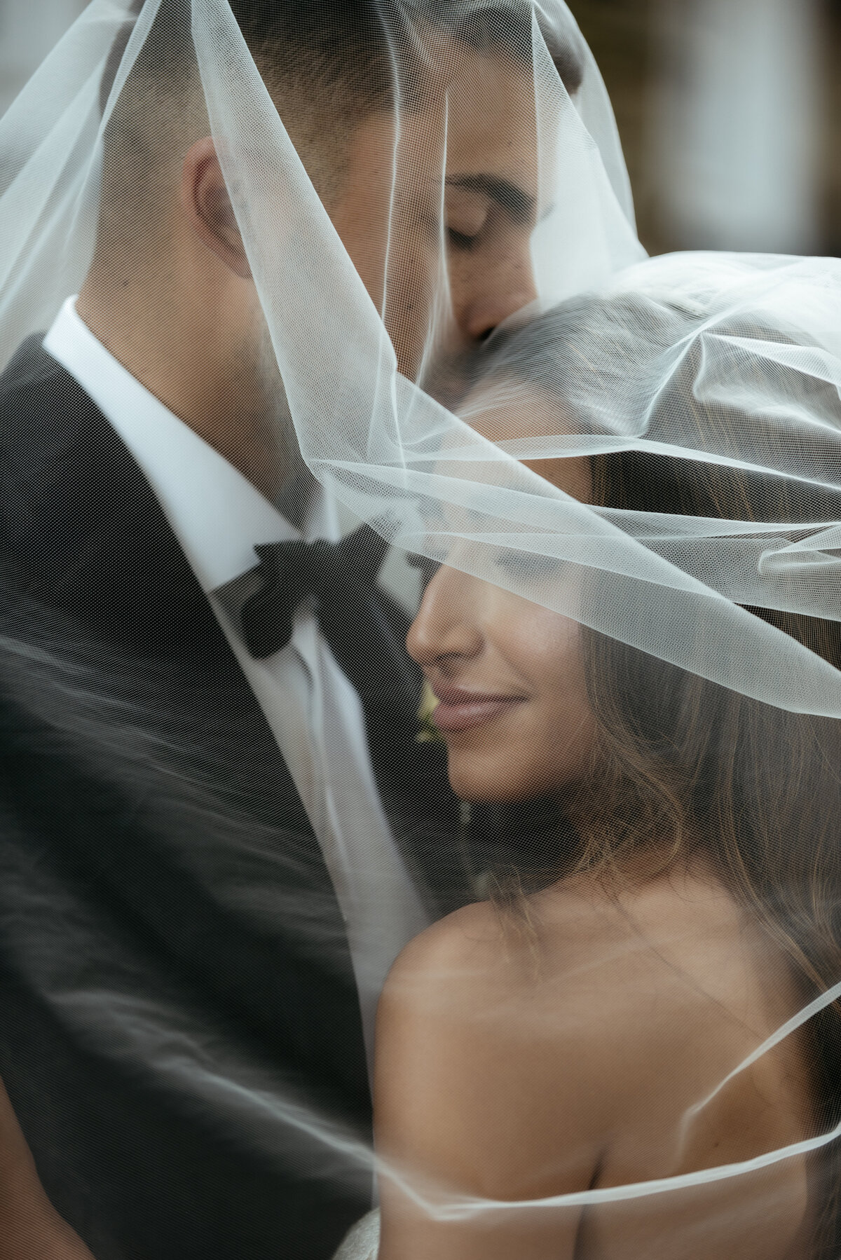 The groom kisses the bride's head as they stand under the veil in moody light.
