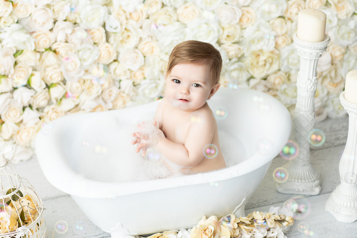 Dallas milestone photographer captures a joyful baby in a white tub surrounded by soft white roses and bubbles, creating a dreamy and elegant bath scene after a cake smash session.