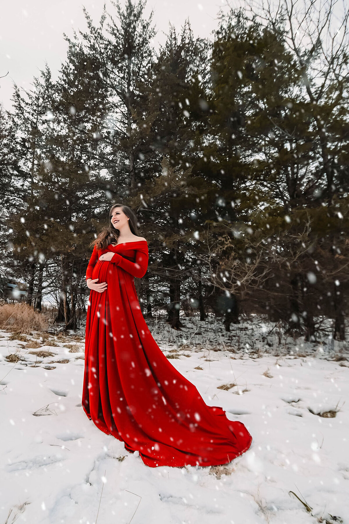 A happy mom to be stands in falling snow in a red maternity gown for her Iowa maternity photographer
